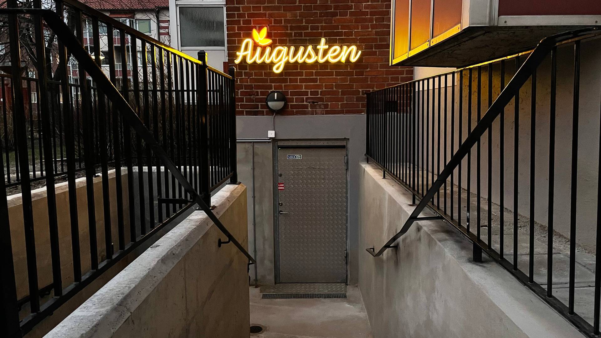 Bilden visar ingången till Augusten som är en trappa ner i en källare. Augusten skylten lyser i neon ovanför dörren.